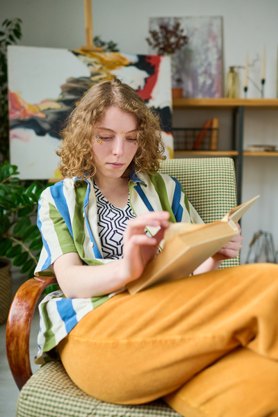A young woman with light curly hair in a bright outfit is reading a book sitting in an armchair with her legs crossed