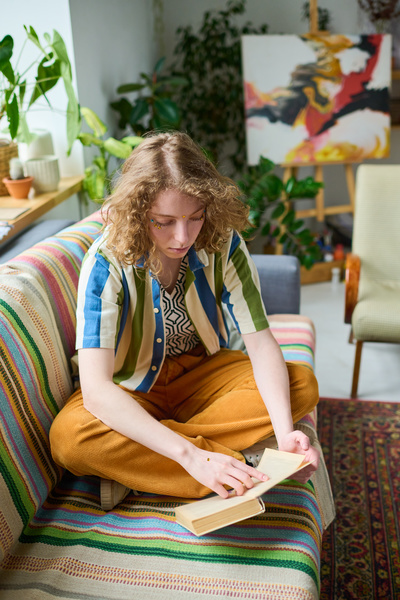 Zoomer girl with light curly hair in bright clothes reads a book sitting in a lotus position on a sofa covered with a striped blanket