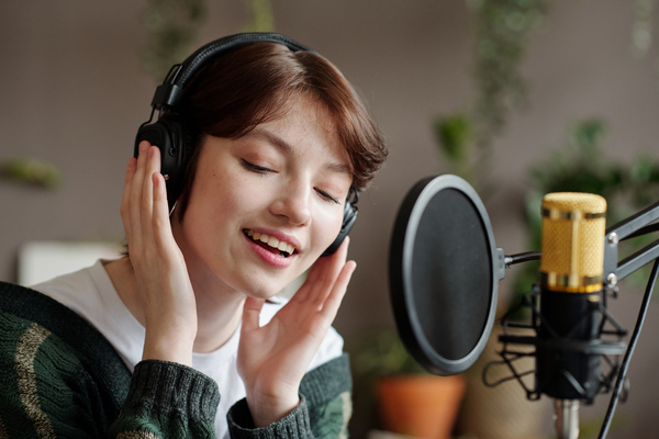A female zoomer with short dark hair in black headphones holding them with both hands sings into a microphone with a pop filter