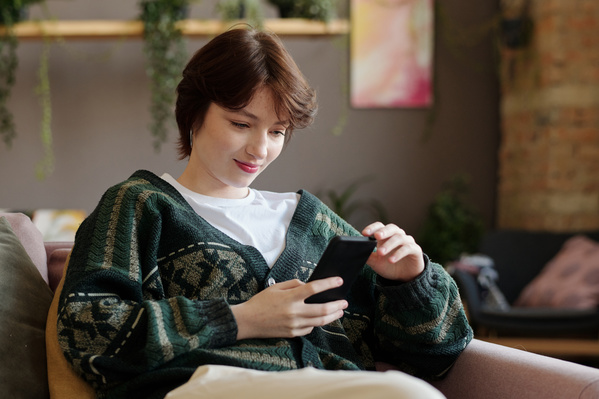 Zoomer girl with a short haircut in a green knitted cardigan is sitting on the couch and smiling looking at her smartphone