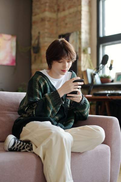 A zoomer girl with short black hair in a dark green knitted cardigan with an ornament and sneakers is sitting on the sofa with her leg bent under her and using her smartphone