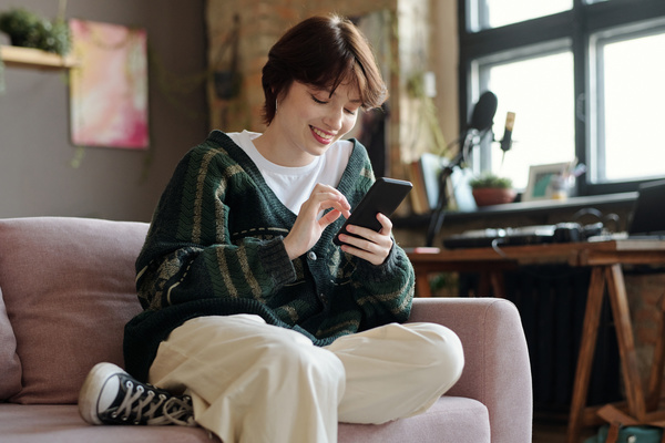 A young woman with short dark hair in a green cardigan and light trousers and sneakers is sitting on the sofa bending her leg under her and smiling using her smartphone