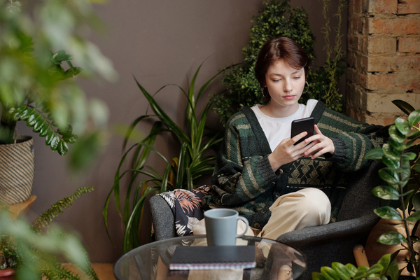 A zoomer woman with short hair in a green jacket and a light T-shirt is sitting on a gray armchair with a smartphone in her hands next to a table in a room with a lot of plants
