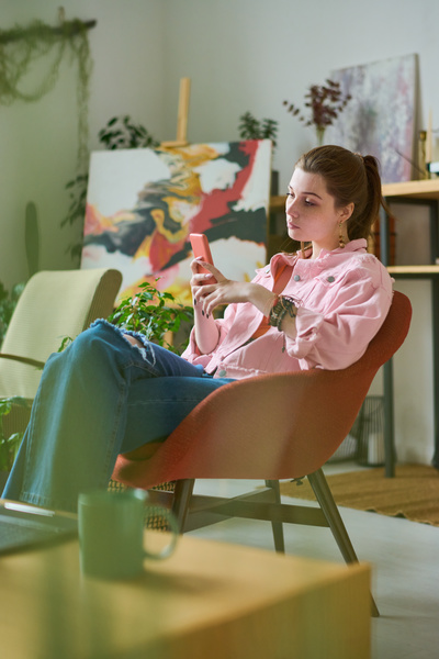 A young woman with her hair in a ponytail in a pink denim jacket and jeans with bracelets on her arm is sitting in a red armchair and looking at her phone