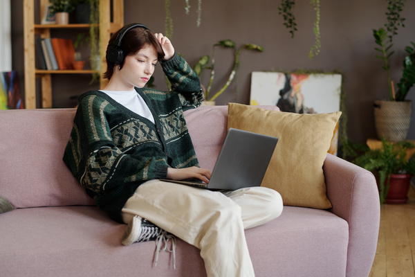 A zoomer woman with short dark hair in black headphones and a dark green cardigan over a white T -shirt looks thoughtfully at a laptop while sitting on a sofa with a pillow bending her leg under her