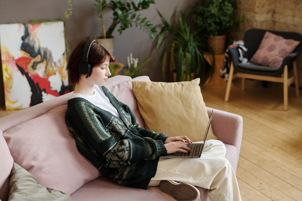 A young student girl with a short haircut wearing black headphones and a green cardigan with a pattern over a white T-shirt is typing on a laptop while sitting on a sofa with pillows with her leg bent under her