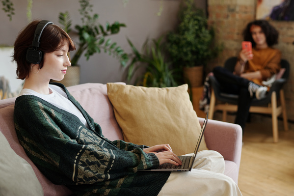 A young girl with dark short hair in headphones and a dark-colored cardigan over a light T-shirt is sitting on a pink sofa with pillows and typing on a laptop in a room with plants