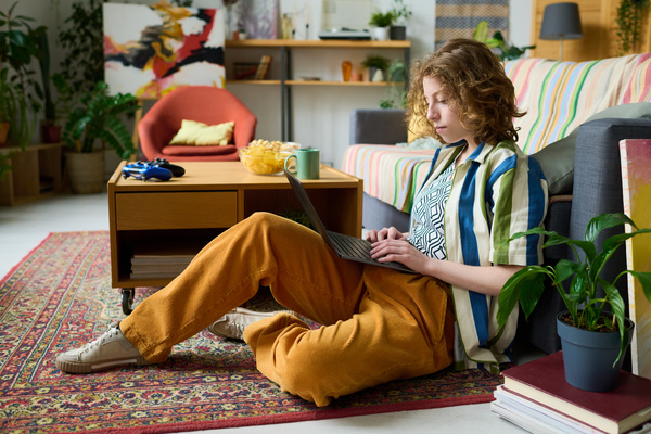 A girl with short curly hair and bright clothes uses a laptop sitting on the carpet with her back to the sofa and bending her legs next to a coffee table with snacks and joysticks on it