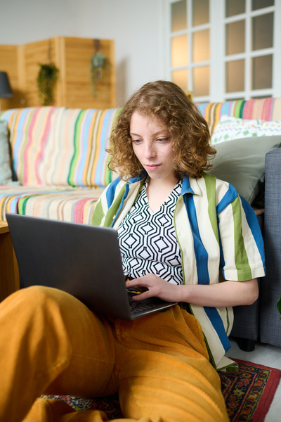 A young girl with light wavy hair and in a bright outfit is typing on a laptop while sitting on the carpet with her back to the sofa