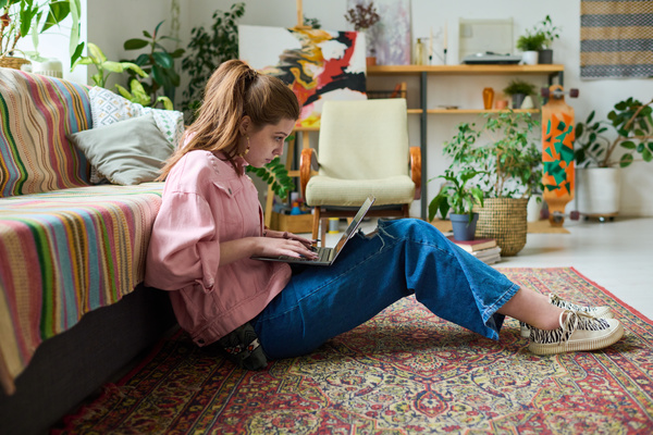 A young woman with gathered hair in a denim pink jacket is sitting on the carpet with her back against the sofa and typing on a laptop