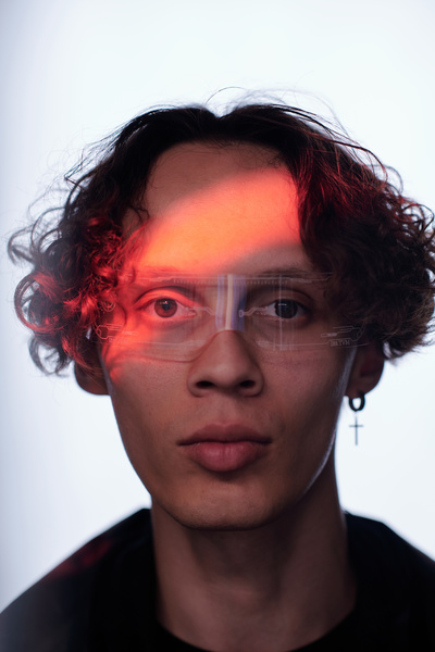A portrait of a man with brown curls with a cross earring in his ear and rimless sunglasses looking straight with a strip of red light on his face