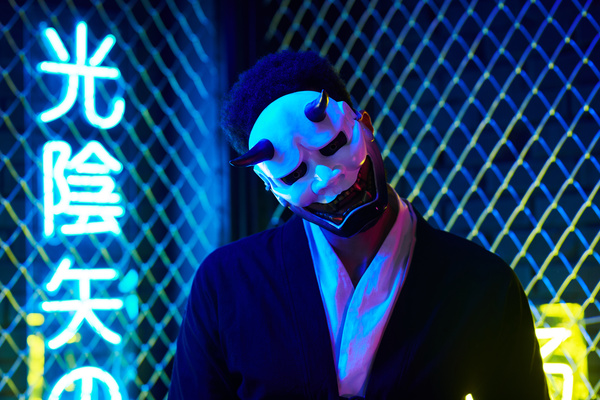 A man with dark hair and wearing a black and white suit stands with his head bent down with a white mask of Hannya with a grimace and horns on his face in blue light against a background of fence and neon hieroglyphics