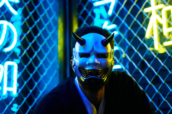 A young man in a white mask of Hannya with fangs and dark clothes with an iron fence and glowing neon yellow and blue symbols behind