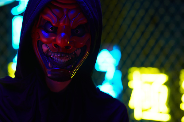 A close-up of a man dressed in black clothes with a hood and a mask of an evil laughing Tengu looking straight ahead and grinning standing against the background of a metal grid behind which bright blue and yellow neon signs
