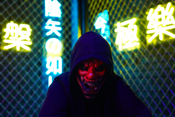 A hooded black-clad person in a mask of a laughing Tengu stands with his head down and yells against a metal mesh with bright blue and yellow neon hieroglyphs behind it