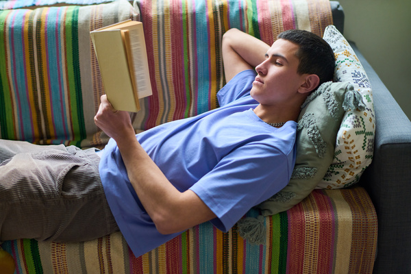 A guy with short black hair in a blue T-shirt and corduroy gray pants is reading a book lying on the couch with one hand behind his head