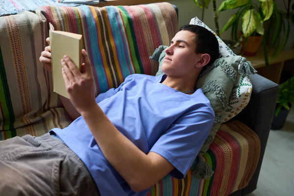 A guy with dark hair in a blue T-shirt and corduroy gray pants is reading a book lying on the sofa with a striped plaid
