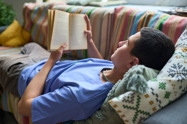 A man with dark hair with a chain around his neck in a blue T-shirt reads a book holding it with both hands and lying on the couch