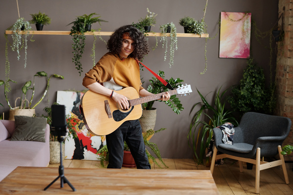 A young curly-haired guy in a sweatshirt stands with a guitar and records a video on a phone that stands on a tripod on a table in front of him in a room with plants