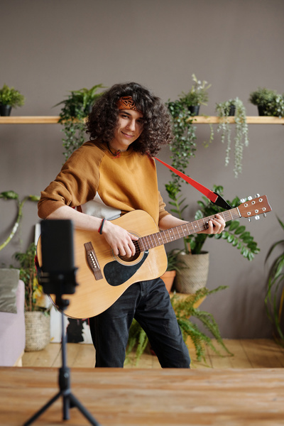 A young man with dark curly hair wearing a bandana on his head in a sweatshirt stands taking a video with an acoustic guitar on a phone standing on a tripod