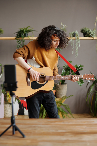 Curly-haired guy with a bandana plays guitar while shooting a video on a phone that stands on a table on a tripod