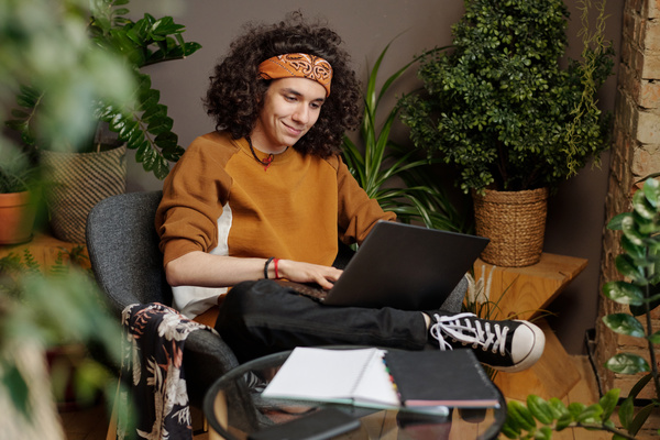 A guy with black curls in a bandana is sitting on a chair with his legs crossed with a laptop on his lap and smiling typing a message