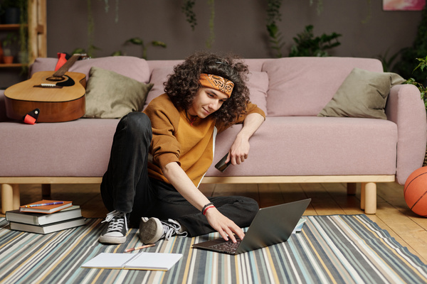 A zoomer guy with curly dark hair is sitting on a striped carpet leaning on the sofa behind with a phone in one hand typing with the other hand in a laptop standing next to him