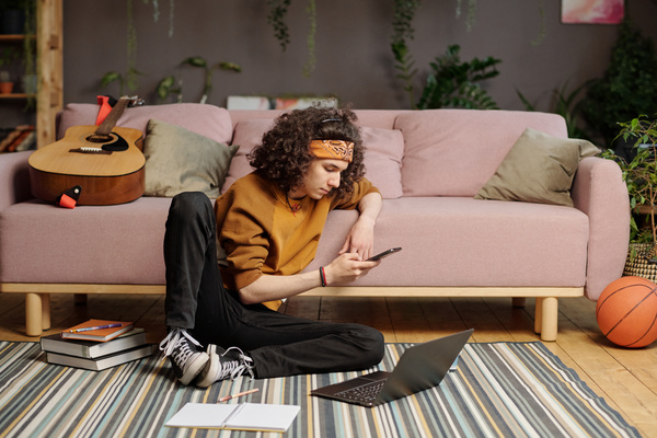 A male zoomer with curly hair is sitting on a striped carpet leaning on a sofa behind with a laptop surrounded by books and using a phone