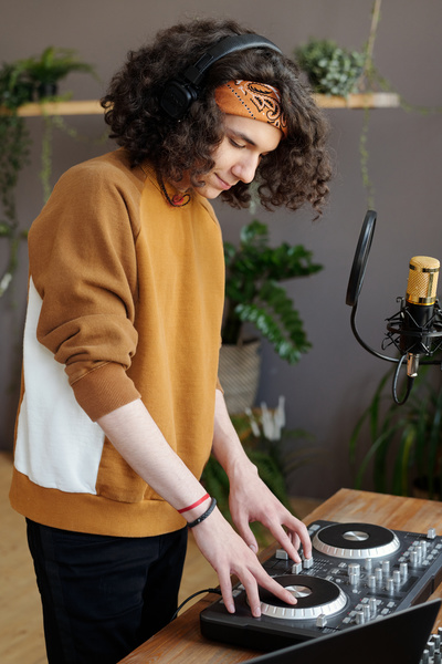 A young man with curly dark hair in a bandana and black headphones is standing at a table on which there is a DJ console and spinning discs at a microphone with a pop filter