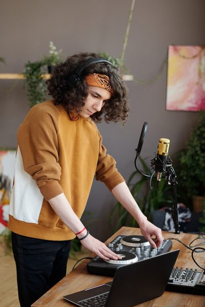A guy with curly black hair wearing a headband and black headphones is standing at a table with a DJ console on it and using it