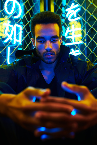 A close-up of a guy with a short afro in a black jacket holding a phone in his hands and sitting in the corner of a fence made of metal mesh behind which neon hieroglyphs hang