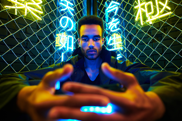 A close-up of a guy with dark curly hair in a black outfit holding a source of bright blue light in his hands and being in the corner of a fence made of metal mesh behind which neon hieroglyphs are