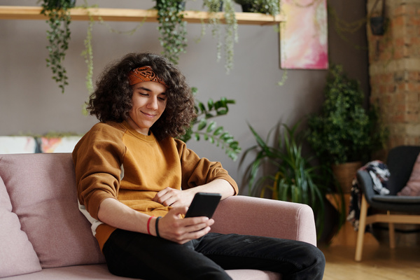 A man with curly hair in a mustard-colored bandana and sweatshirt is sitting on a pink sofa in a bright room with green plants and smiling surfing social networks on his smartphone