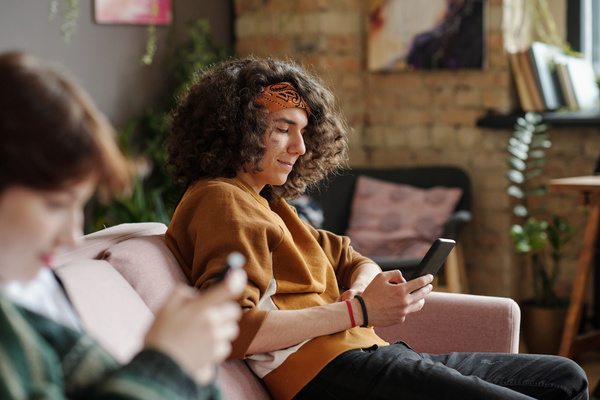 A zoomer guy with a lush hairstyle in a bandana sits on the couch with a phone in his hand and smiling looks at its screen