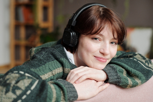 A young girl with short dark hair wearing headphones and a dark green knitted sweater with patterns is sitting on the sofa with her head on her hands and smiling looking in front of her