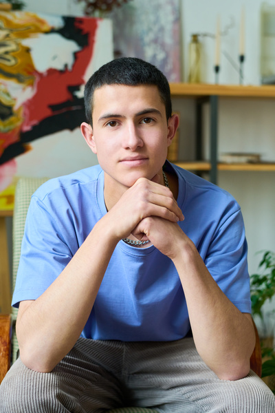 A handsome young man with black hair and brown eyes dressed in a blue T-shirt and corduroy gray trousers sits with his elbows on his knees and looks in front of him