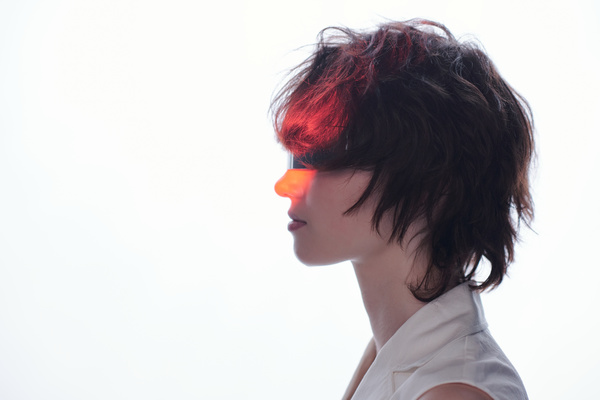 On a white background in the shade the profile of a young woman with a red neon beam on her face with short disheveled brown hair wearing glasses with a silvery lens