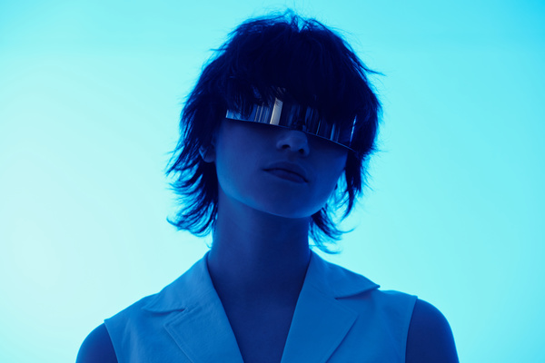 A girl in a futuristic style with short tousled hair wearing glasses with a metal lens dressed in a light blouse with a double-breasted collar stands under neon blue light on a light background