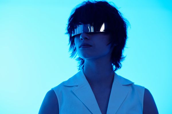 A young woman in a futuristic style with short tousled hair and in glasses resembling a metal plate in a white outfit stands with her head turned to the side in a room with blue lighting