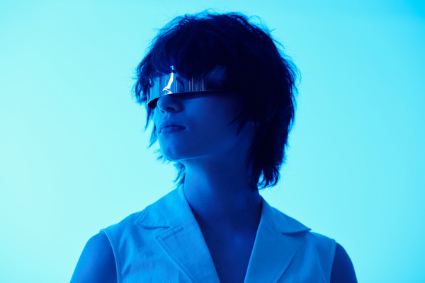 A futuristic looking woman with short hair and in silver-colored glasses turned her head while standing under a neon blue light on a white background