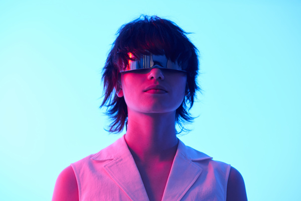 A girl with short curly hair wearing glasses resembling a metal plate stands slightly raising her head under the light of red and blue neon on a light background