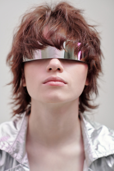 Full-face of of a fresh-looking woman on a light-colored background with carelessly styled short brown hair wearing silver-colored clothing with a collar and metallic glasses with a pink glow