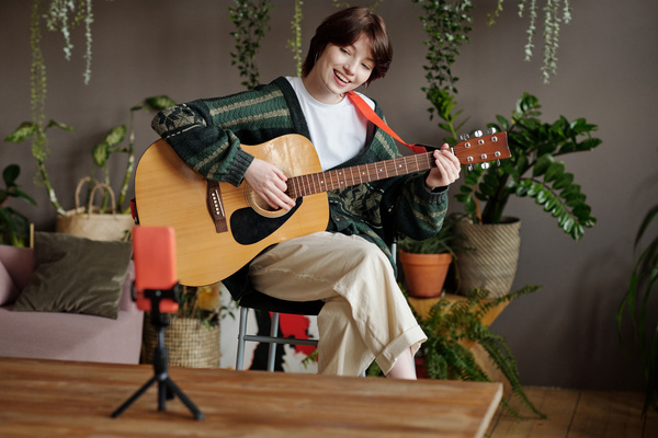 A young woman with short black hair in a cardigan and light trousers crossing her legs and smiling to the teeth plays an acoustic guitar and sings while filming herself on an orange phone on a tripod