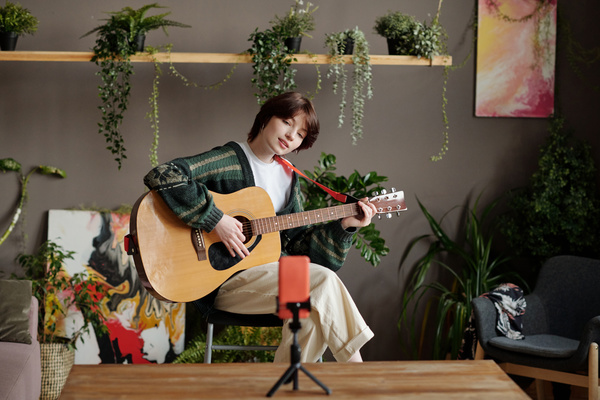 A zoomer girl with a short haircut tilts her head and shoots herself on the phone playing an acoustic guitar in a room with green plants