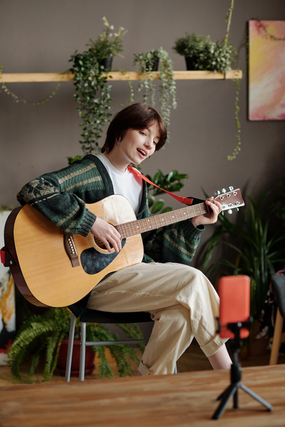 A zoomer woman with a pixie haircut in a green patterned cardigan and beige trousers sitting on a chair sings while playing the guitar