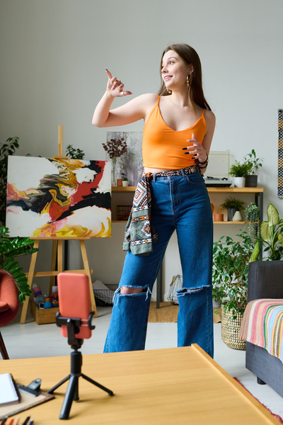 A girl with long loose hair in a bright top and jeans with a patterned belt in a bright room moves shooting video on a phone standing on a tripod