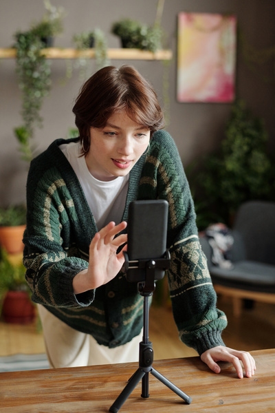 A girl with short dark hair in a dark green cardigan over a light T-shirt leaning her hand on a table on which there is a phone on a tripod is going to take a selfie