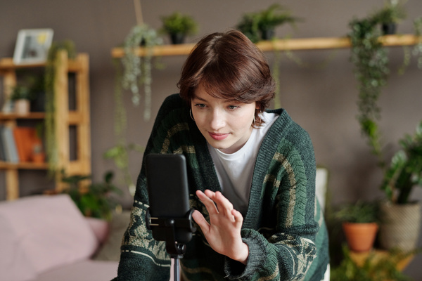 A young woman with a pixie haircut in a patterned cardigan standing next to a phone on a tripod is going to take a photo of herself
