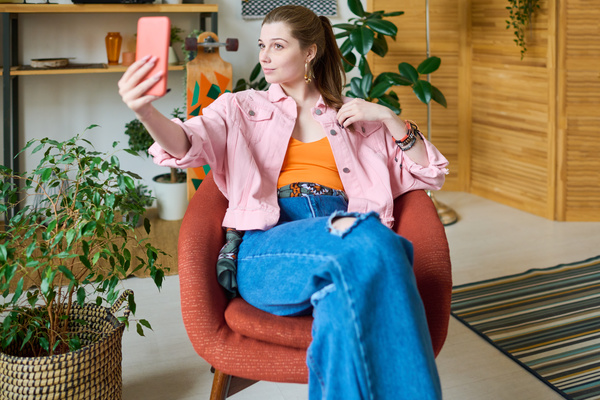 A young woman with a ponytail in bright clothes sits on a red chair in a bright room and takes a selfie on her phone