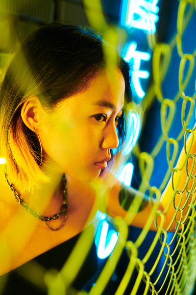 Young girl with short dyed hair at the tips and a necklace dressed in a black top stands behind a metal mesh fence lit with yellow light